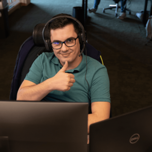 Portrait of persona: Wojtek. He is sitting in front of the computer. He is looking into a camera and shows thumbs up gesture.